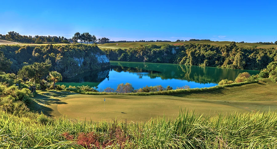 Golf putting green with a flag beside a turquoise lake surrounded by limestone cliffs, dense green trees, and rolling fairways under a clear blue sky.