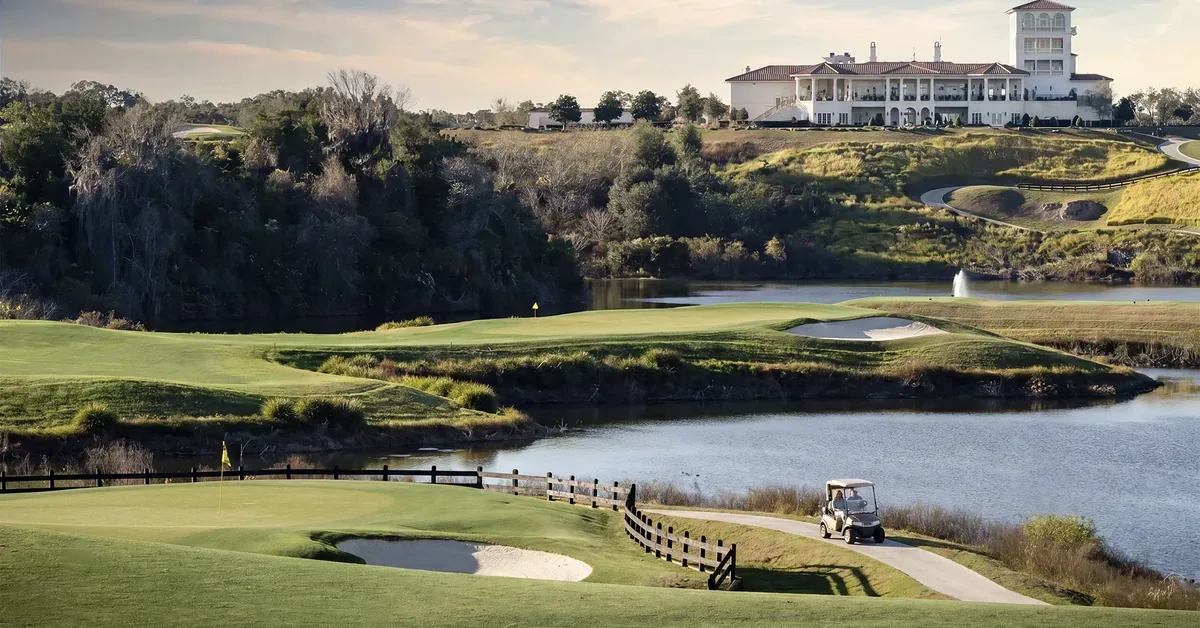 Scenic golf course with manicured fairways, sand bunkers, and a serene lake leads to a grand clubhouse estate perched on a hill.