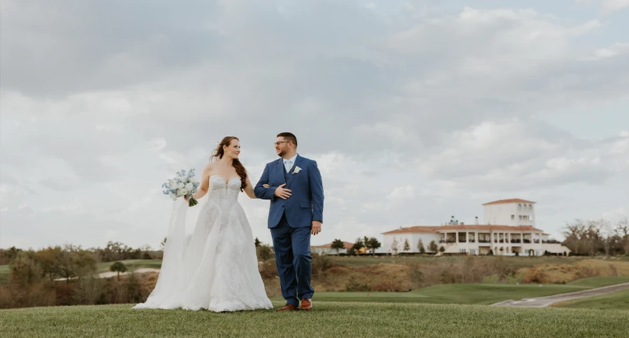 Bride and groom walking across the golf course with The Club at Iron Lake clubhouse in the background.