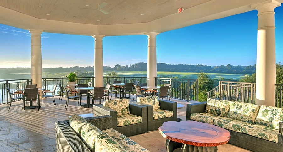 Covered clubhouse terrace with white columns, patterned green-and-cream sofas and chairs, a live-edge wood table, and dining tables overlooking a golf course under a blue sky.