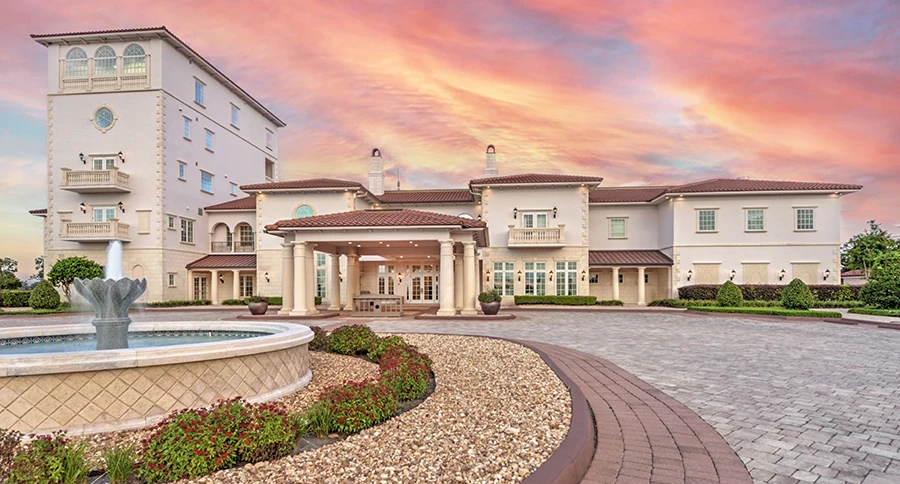 Cream clubhouse with a red tile roof, tower, and columned entrance beside a stone fountain, flower beds, and a brick paver driveway under a pink and orange sunset sky.