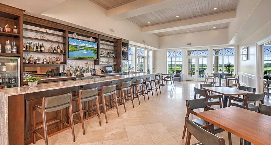 Clubhouse bar with a long stone countertop, wooden barstools with green cushions, shelves of liquor bottles, and a TV, beside a bright dining area with wood tables and glass doors to the golf course.