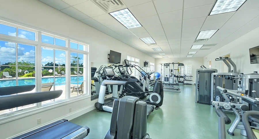 Bright fitness center with treadmills, elliptical machines, and weight machines on a green floor, with large windows overlooking an outdoor swimming pool and lounge chairs.