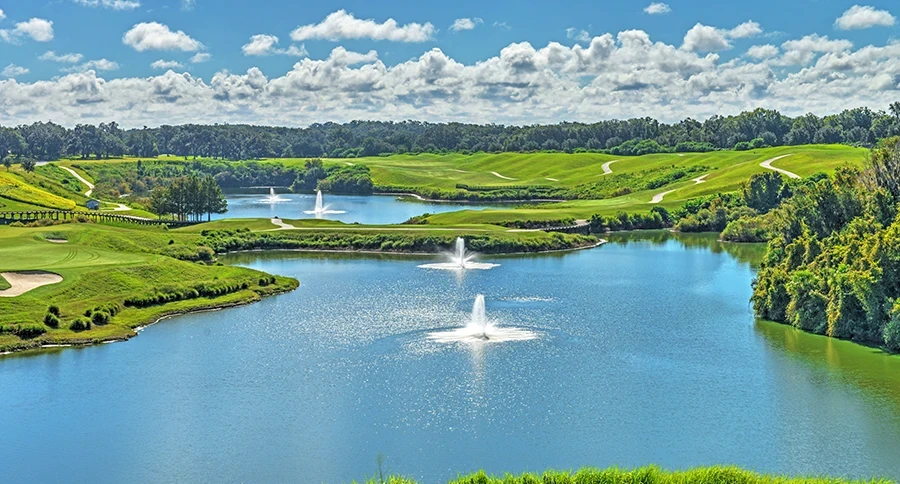 Blue lake with three white fountains surrounded by green rolling golf fairways, a sand bunker, and dense trees under a blue sky with white clouds.