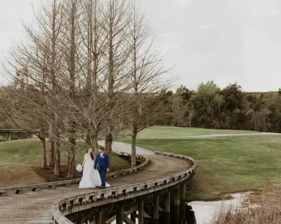 A bride and groom walk hand-in-hand along a curved wooden boardwalk overlooking a lush golf course landscape with a serene pond.