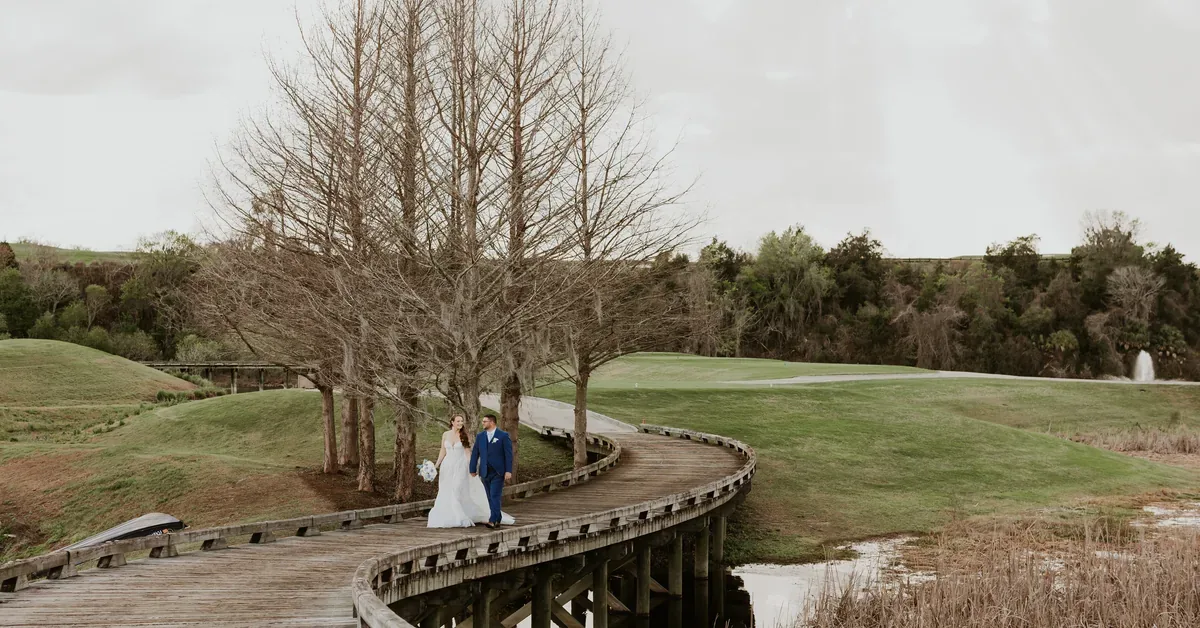 A bride and groom walk hand-in-hand along a curved wooden boardwalk overlooking a lush golf course landscape with a serene pond.