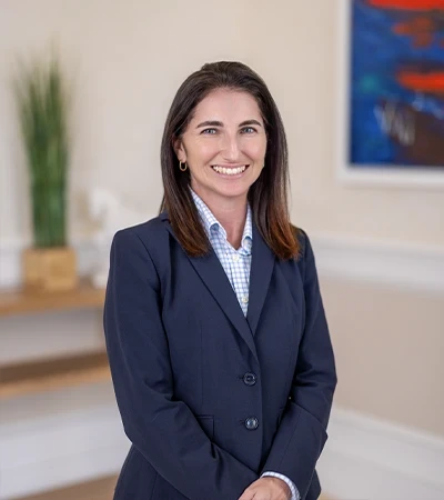 Headshot of Chelsea Addy, a smiling woman with shoulder-length brown hair wearing a navy blazer over a blue checked collared shirt, standing indoors near a potted plant and a red-and-blue abstract painting.