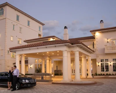Man and woman in evening attire standing beside a black luxury car under a lit column entrance of a cream-colored Mediterranean clubhouse at dusk.
