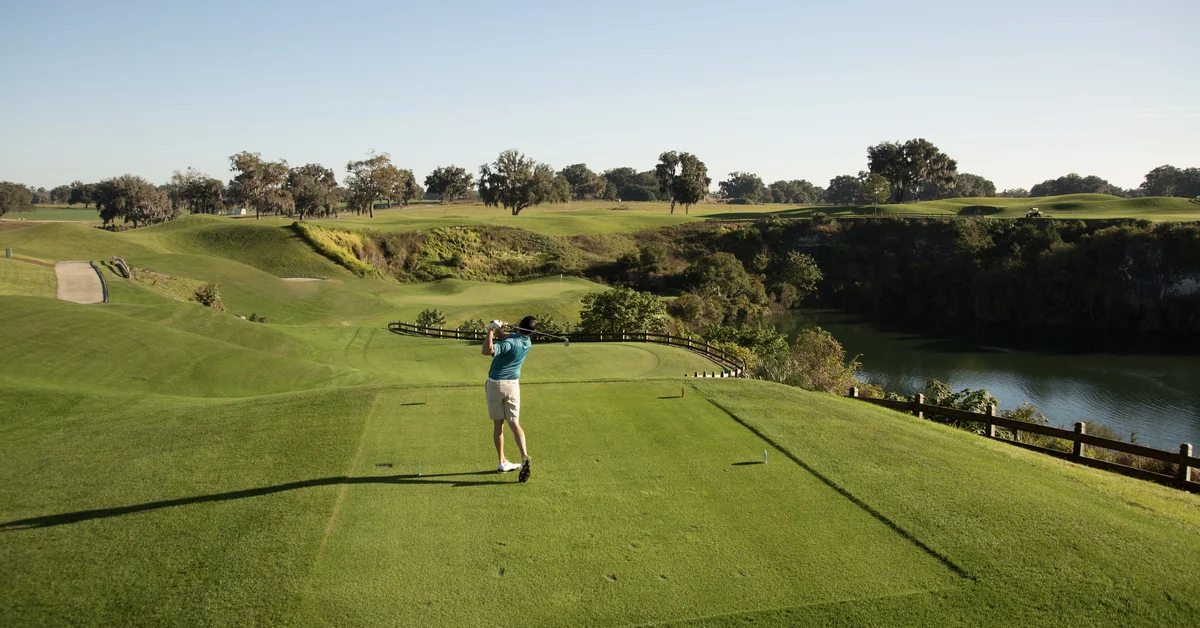 Golfer finishing a swing on a tee box overlooking rolling fairways, a rock quarry, and a lake, with oak trees in the background.