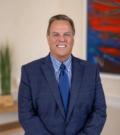 Headshot of Stephen McLean, a smiling man with short gray hair wearing a blue plaid suit, blue striped shirt, and navy tie, standing indoors near a potted plant and a red-and-blue abstract painting.