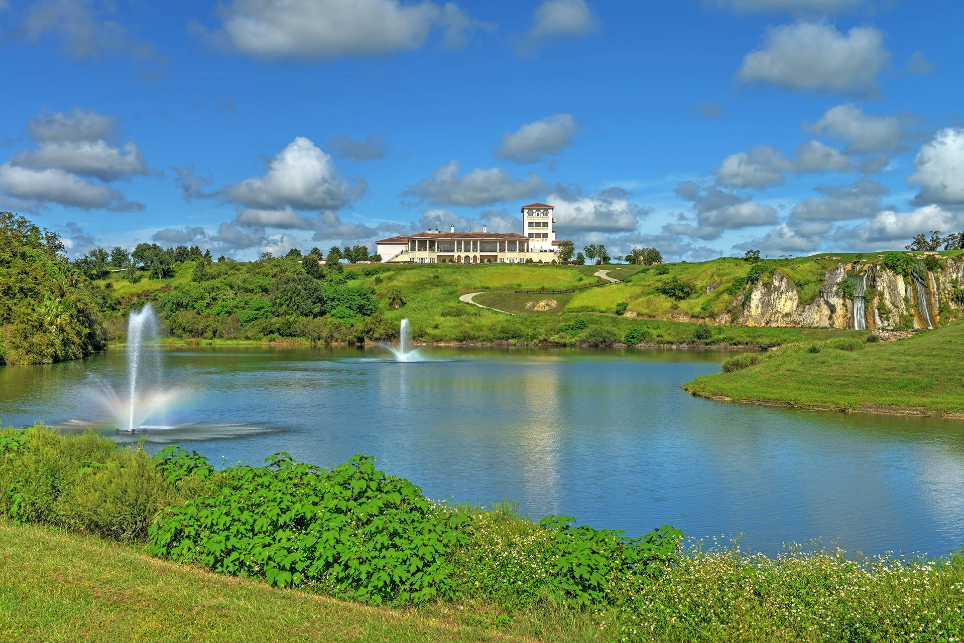 Cream-colored clubhouse with a tower on a green hilltop above a blue lake with two fountains and a limestone cliff with waterfalls, under a blue sky with white clouds.