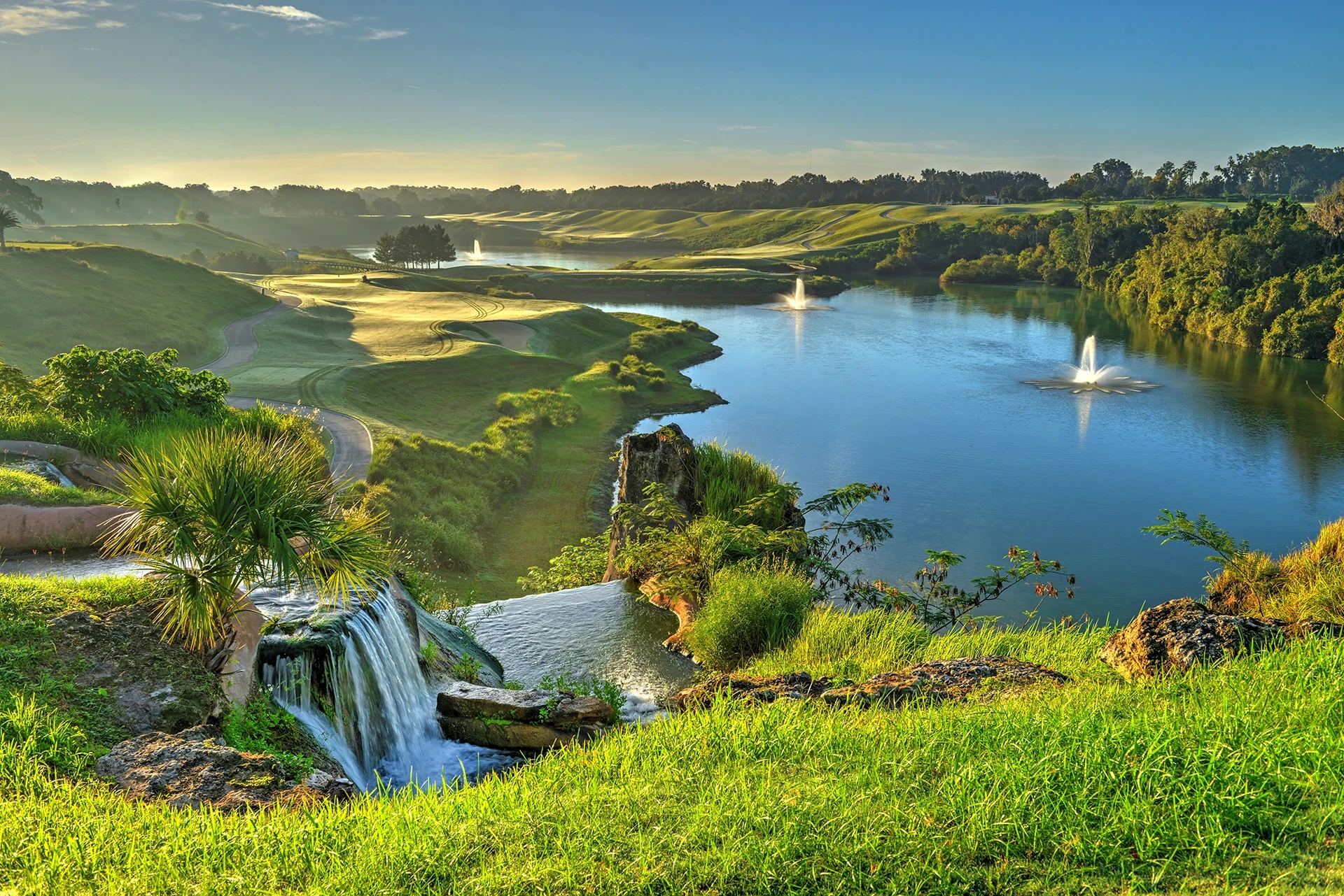 Sunrise view over a rolling golf course fairway and bunker beside a blue lake with two water fountains, with a waterfall over rocks and a palm in the foreground.